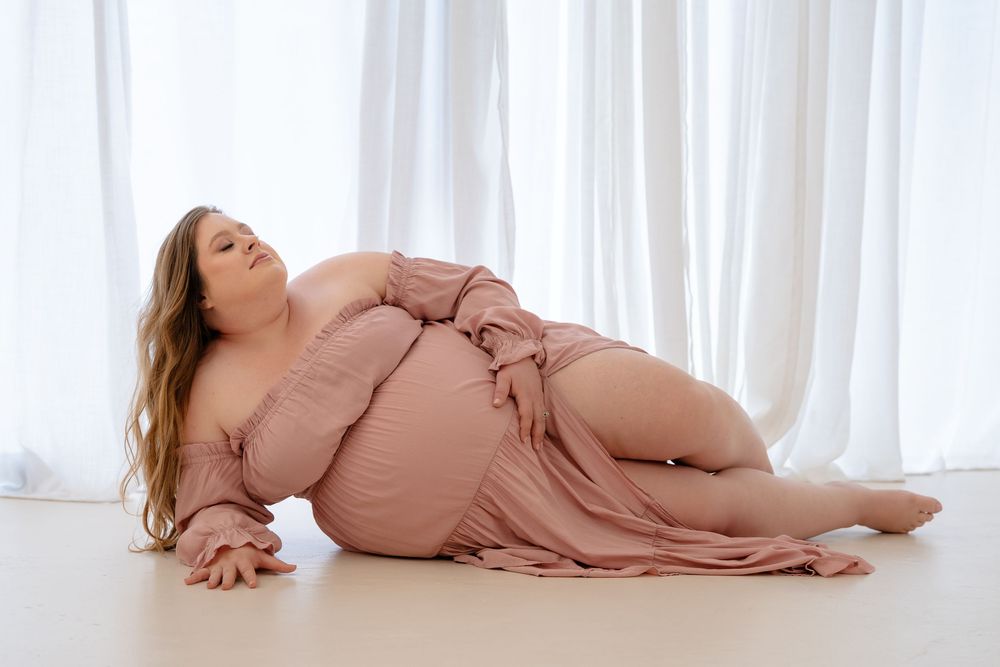 Woman in a dusty rose dress poses gracefully on white floor with sheer curtains in background.