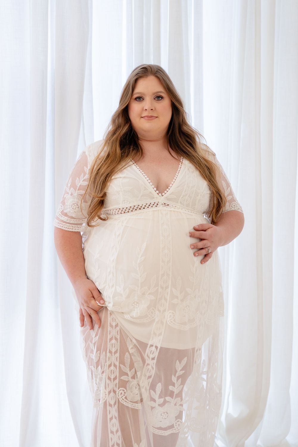 A woman in a cream lace dress poses against a white curtain backdrop in a boudoir style photoshoot.