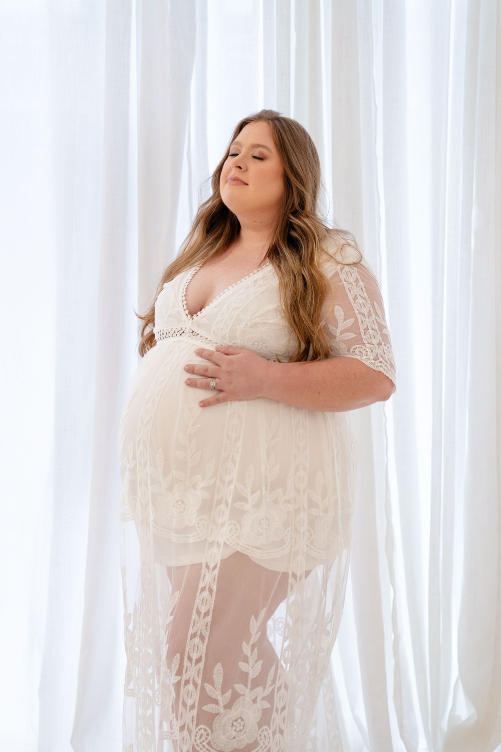 A woman in a flowing white lace maternity dress poses gracefully against sheer curtains in a bright studio setting.