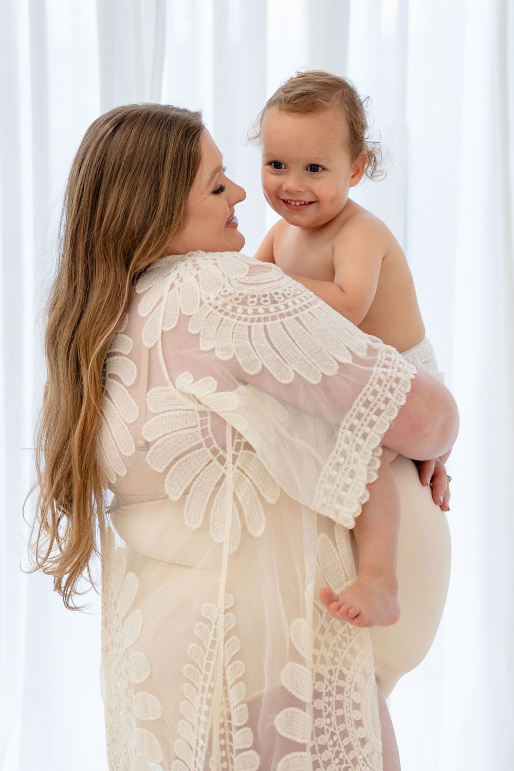 Mother and baby share tender moments in white lace dress against sheer curtain backdrop.
