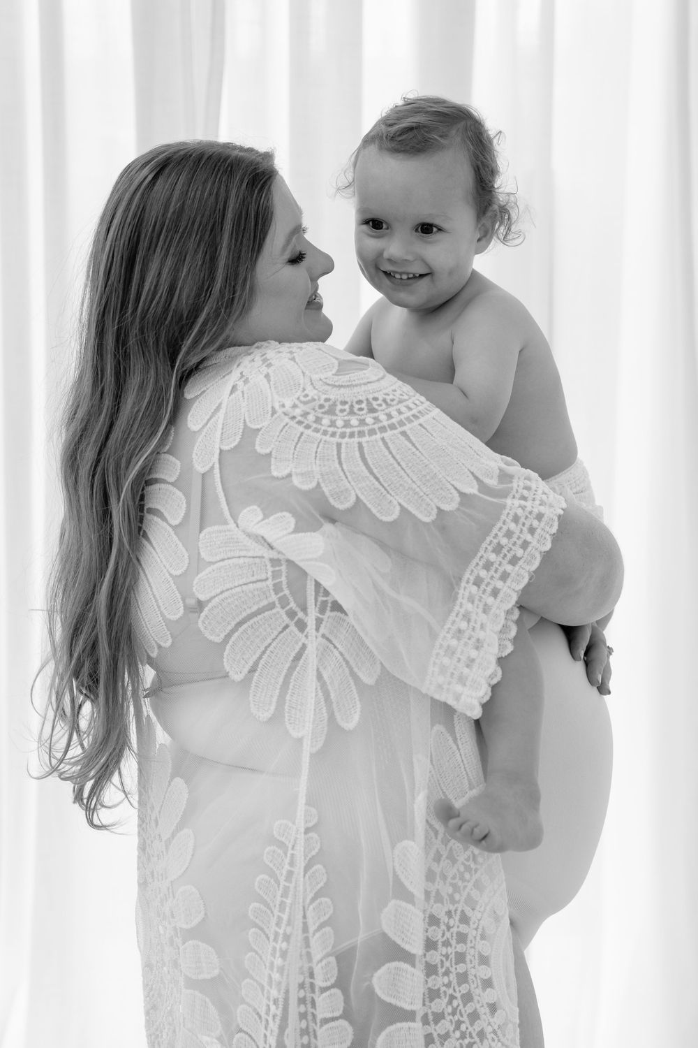 A tender black and white portrait of a parent in a lace dress holding a smiling baby against a light background.