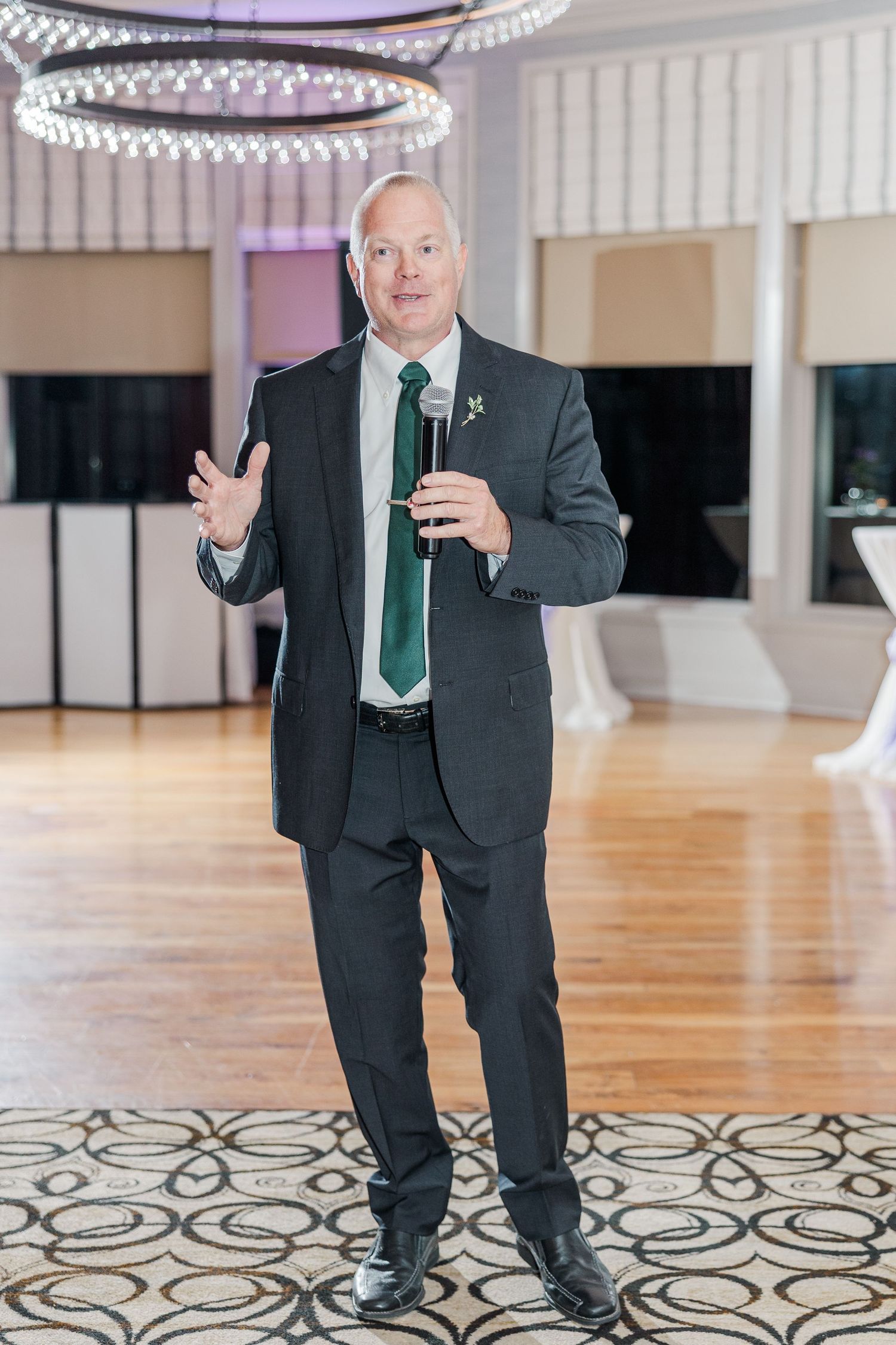 A person in a dark suit and green tie gestures while standing on a patterned ballroom floor.