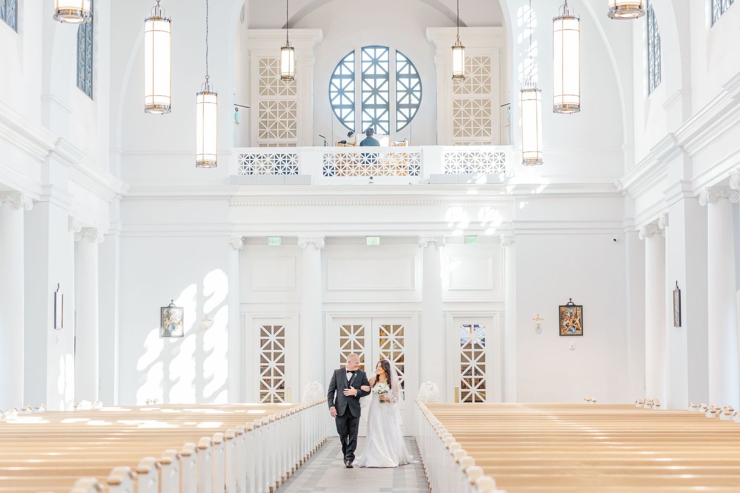 A couple stands at the end of a bright white church aisle with hanging pendant lights and ornate circular window details.