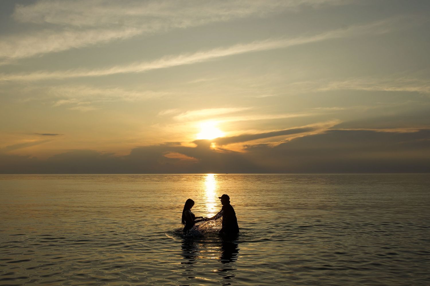 Two silhouettes sit in a small boat on calm waters during a golden sunset over distant mountains.