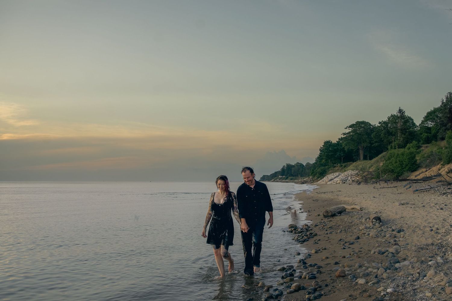 A couple walks along a rocky beach at sunset with trees and coastline in the background.