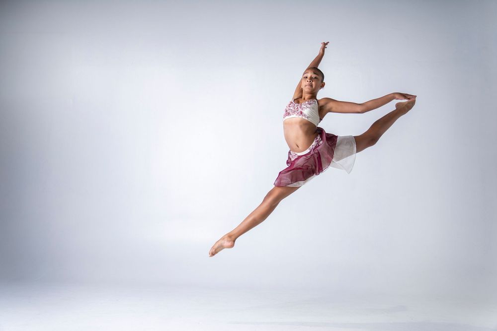 A dancer in pink attire performs a graceful split leap against a light gray studio background.