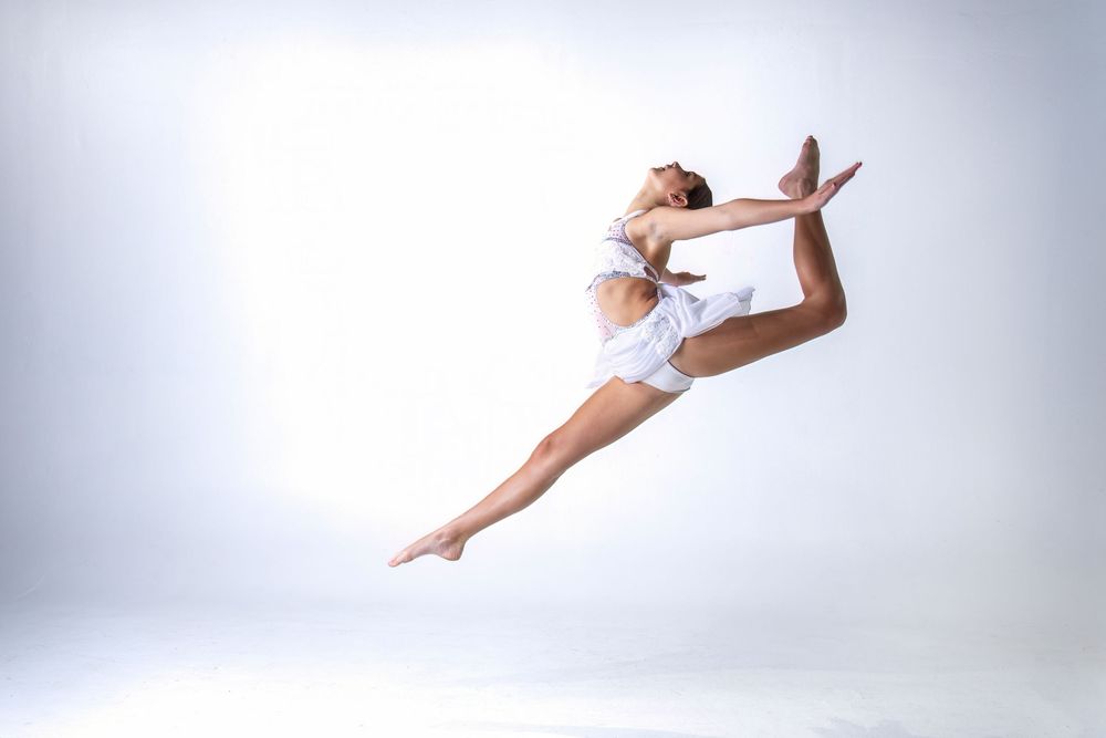 A graceful dancer in white performs a series of dynamic leaps and poses against a light studio backdrop.