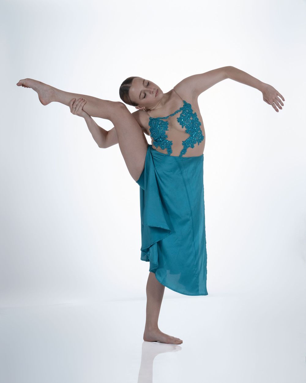 A dancer in turquoise performs a graceful arabesque pose against a white backdrop in a flowing dress.