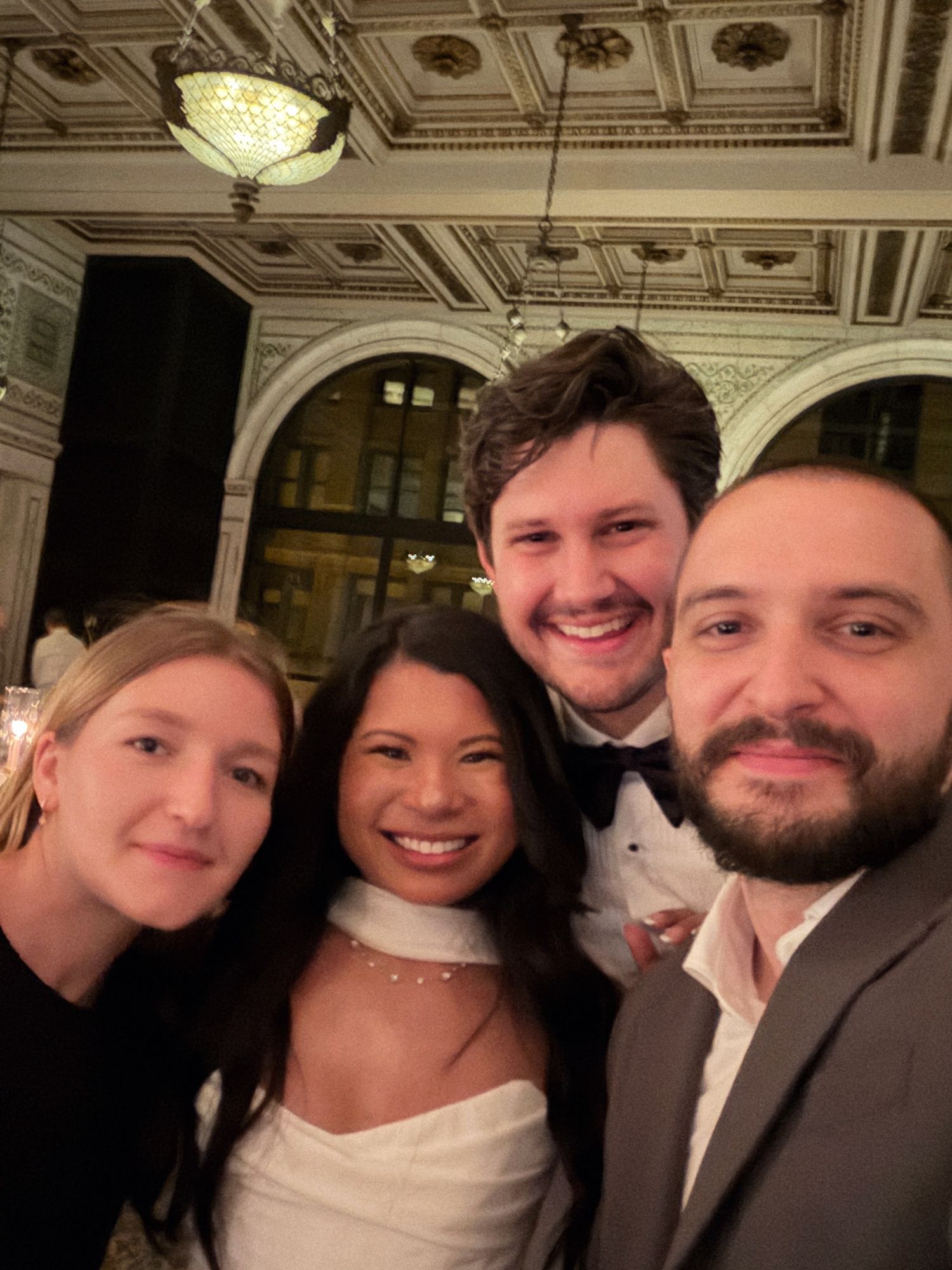 A group of friends pose together at an elegant venue with ornate ceiling and crystal chandelier.