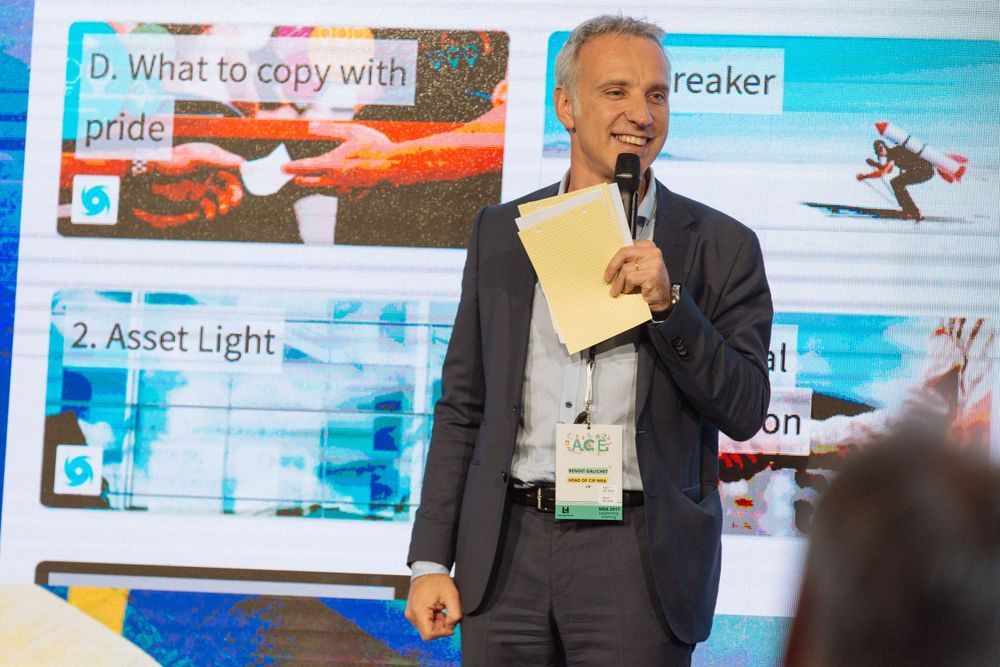 A presenter in a dark suit stands at a podium giving a presentation with digital screens visible in the background.