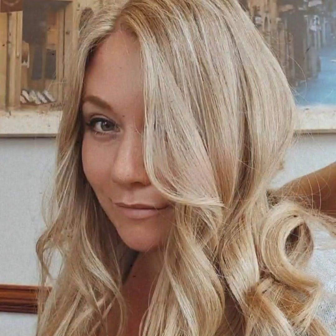 Close-up portrait with long wavy blonde hair styled in loose curls against an interior background.