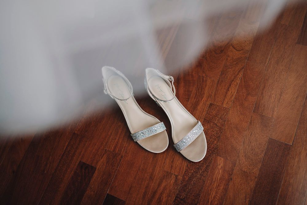 Scarpe da sposa bianche disposte su un pavimento in legno accanto a un tessuto di tulle svolazzante.
