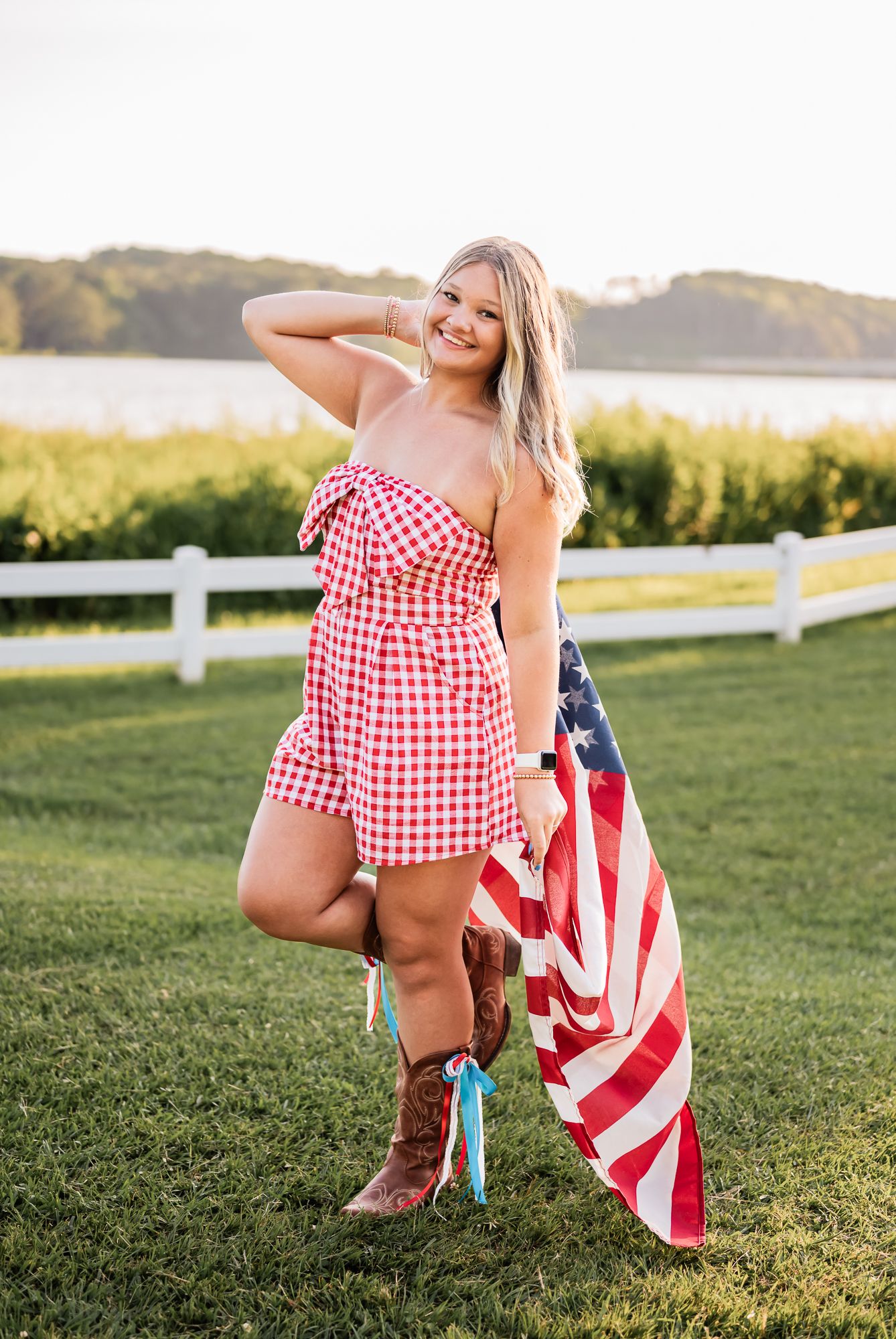 A person in cowboy boots and a red gingham dress holds an American flag in a grassy field at sunset.