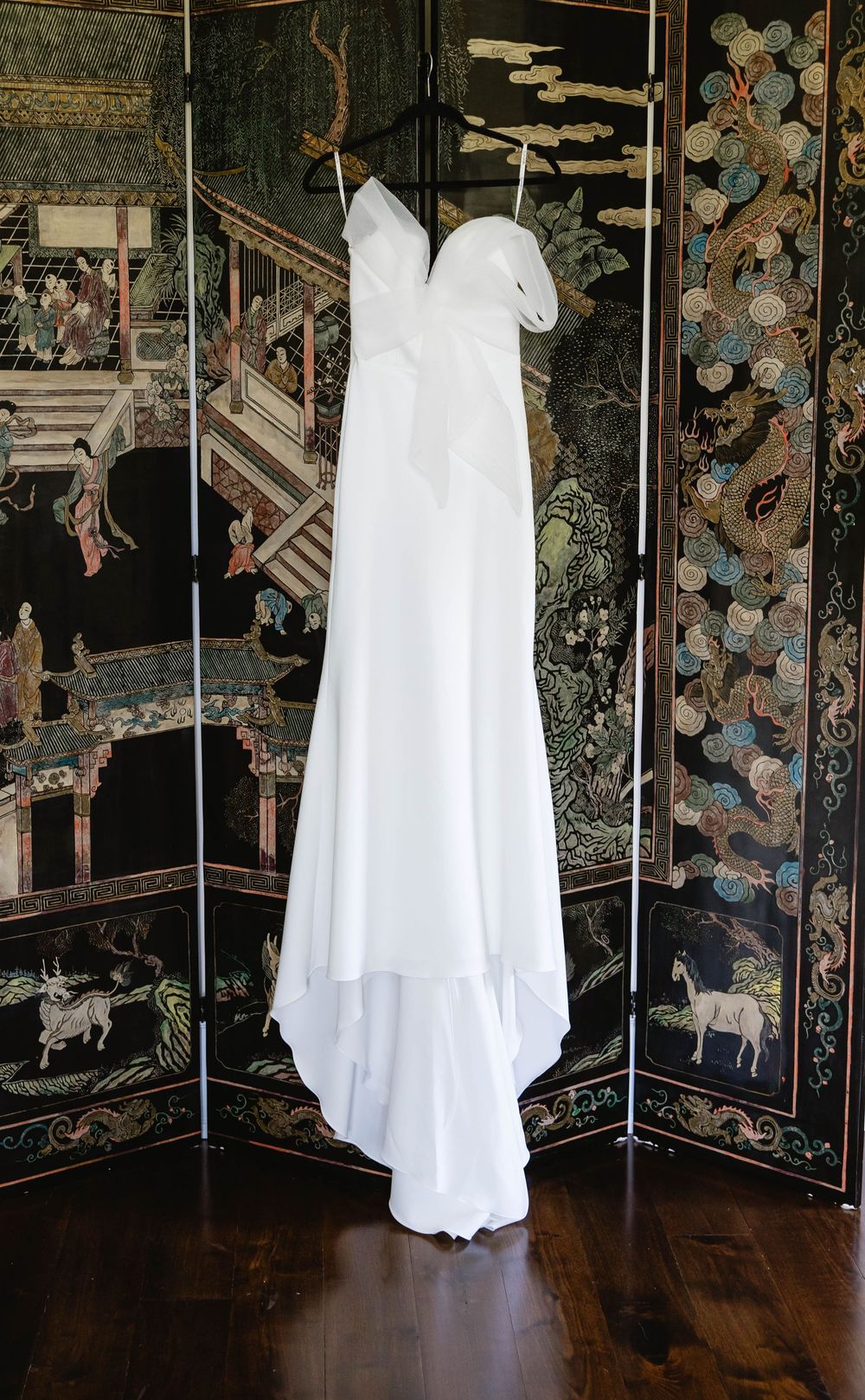 White wedding dress hanging elegantly against a dark decorative wall with floral patterns. Appleford Estate. Villanova, PA.