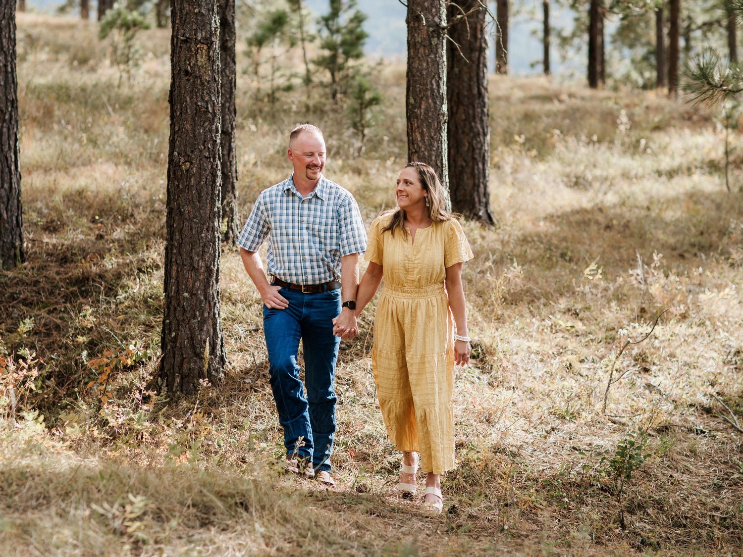 Couple walking hand in hand through sunlit pine forest in casual attire.