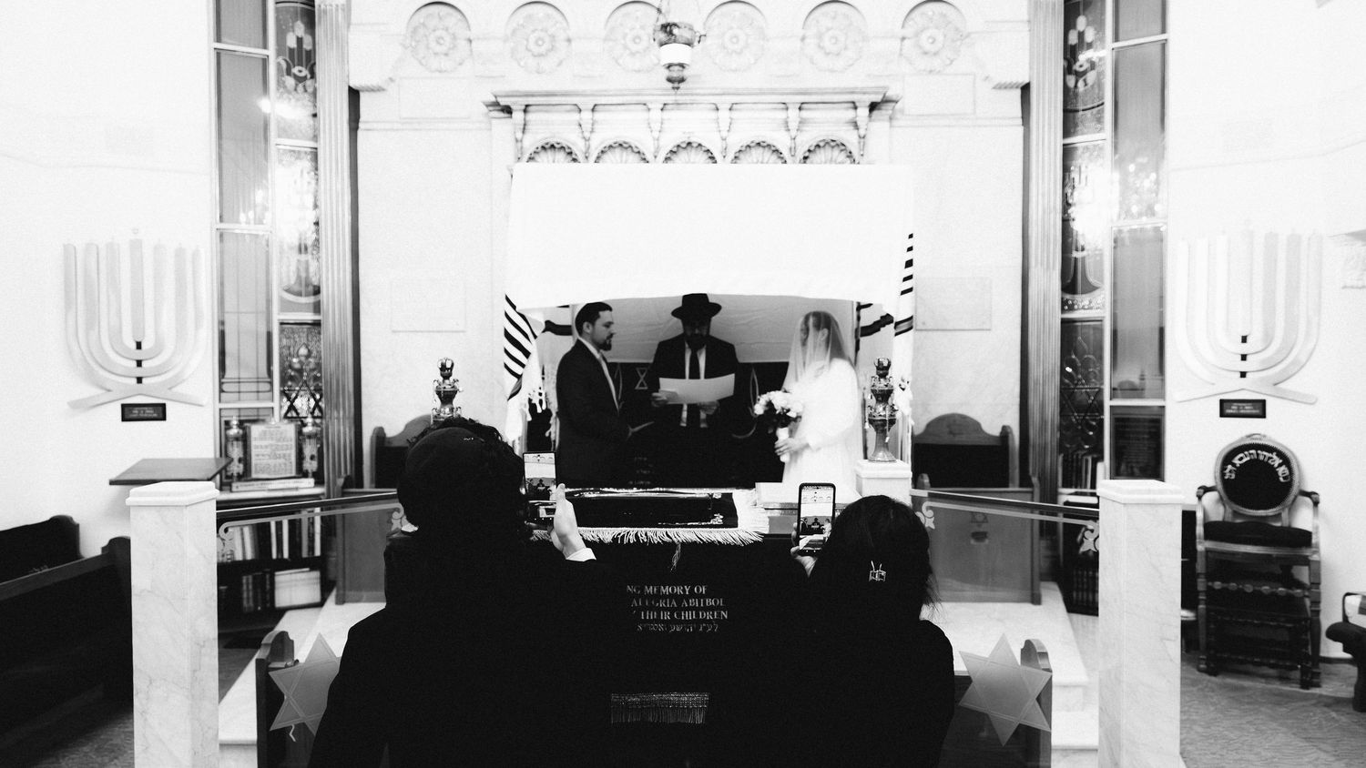 Black and white photo of a Jewish wedding ceremony at a synagogue altar with guests seated in the foreground.