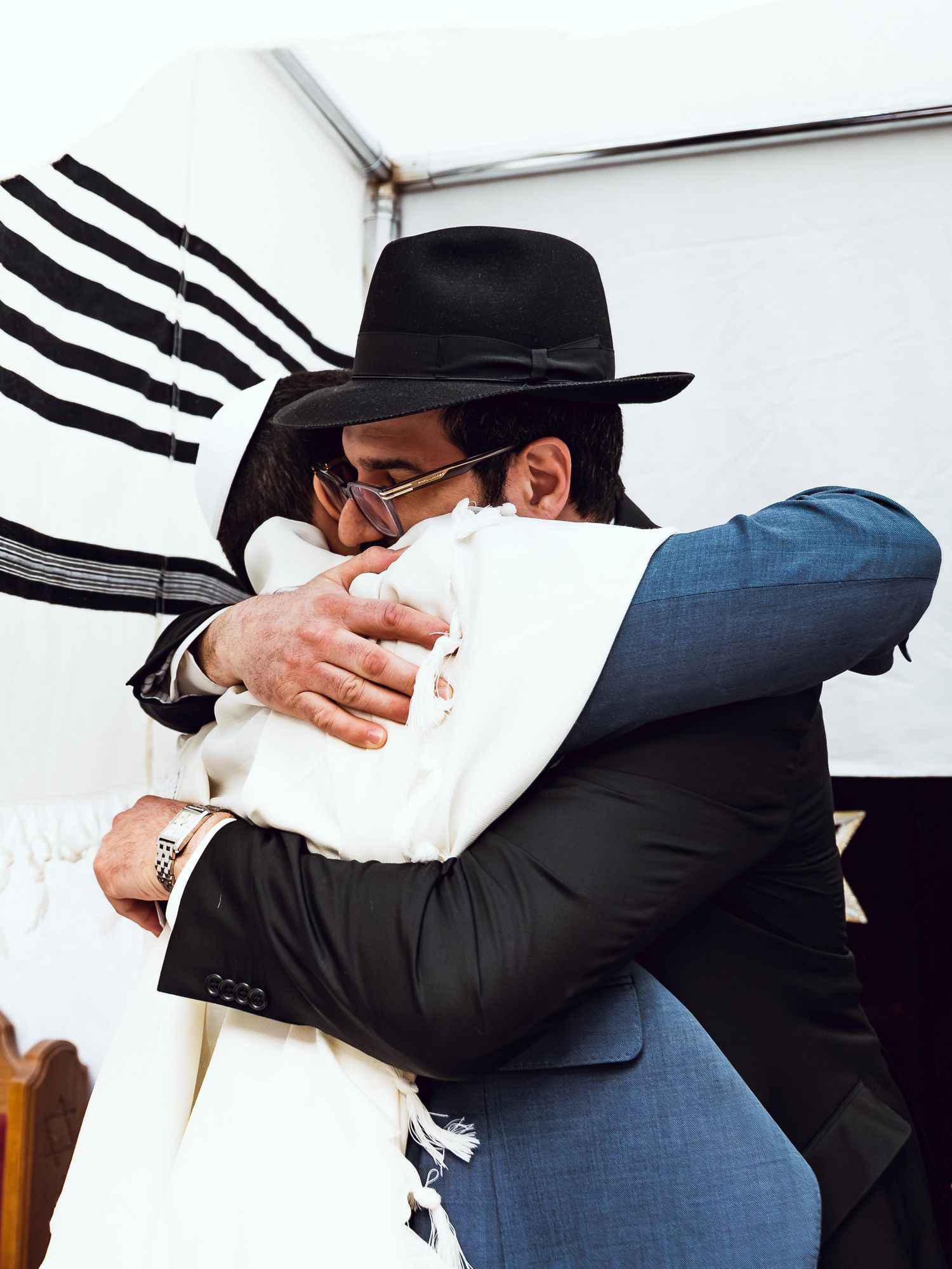 Two men in formal attire share a warm embrace at a Jewish ceremony, one wearing a traditional black fedora hat.