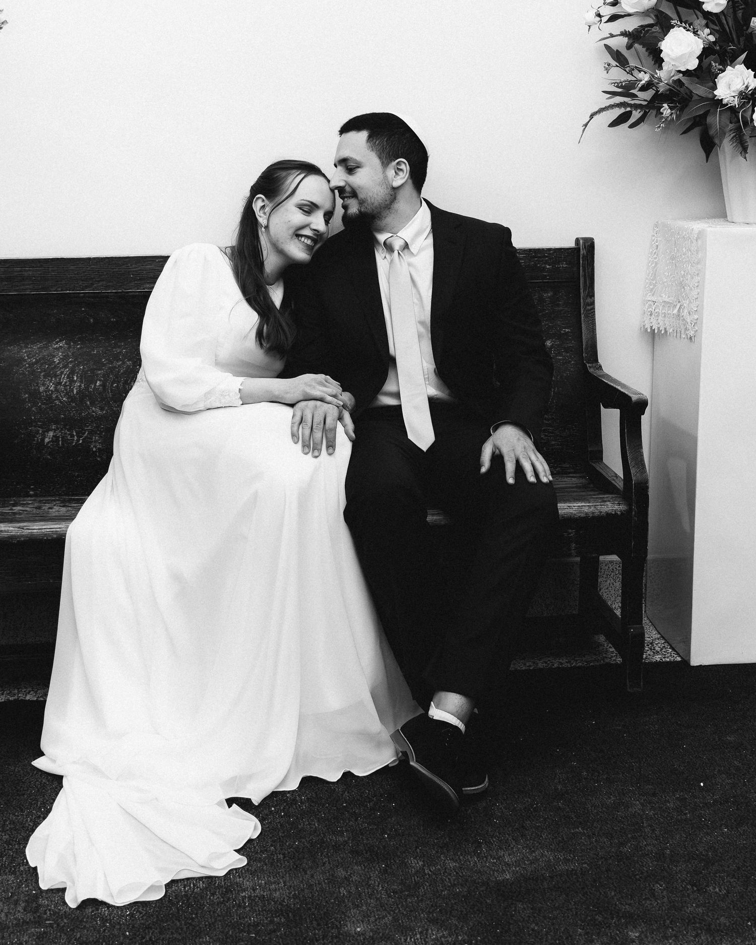 Black and white wedding photo of bride in white gown and groom in suit sitting on bench, sharing a romantic moment.