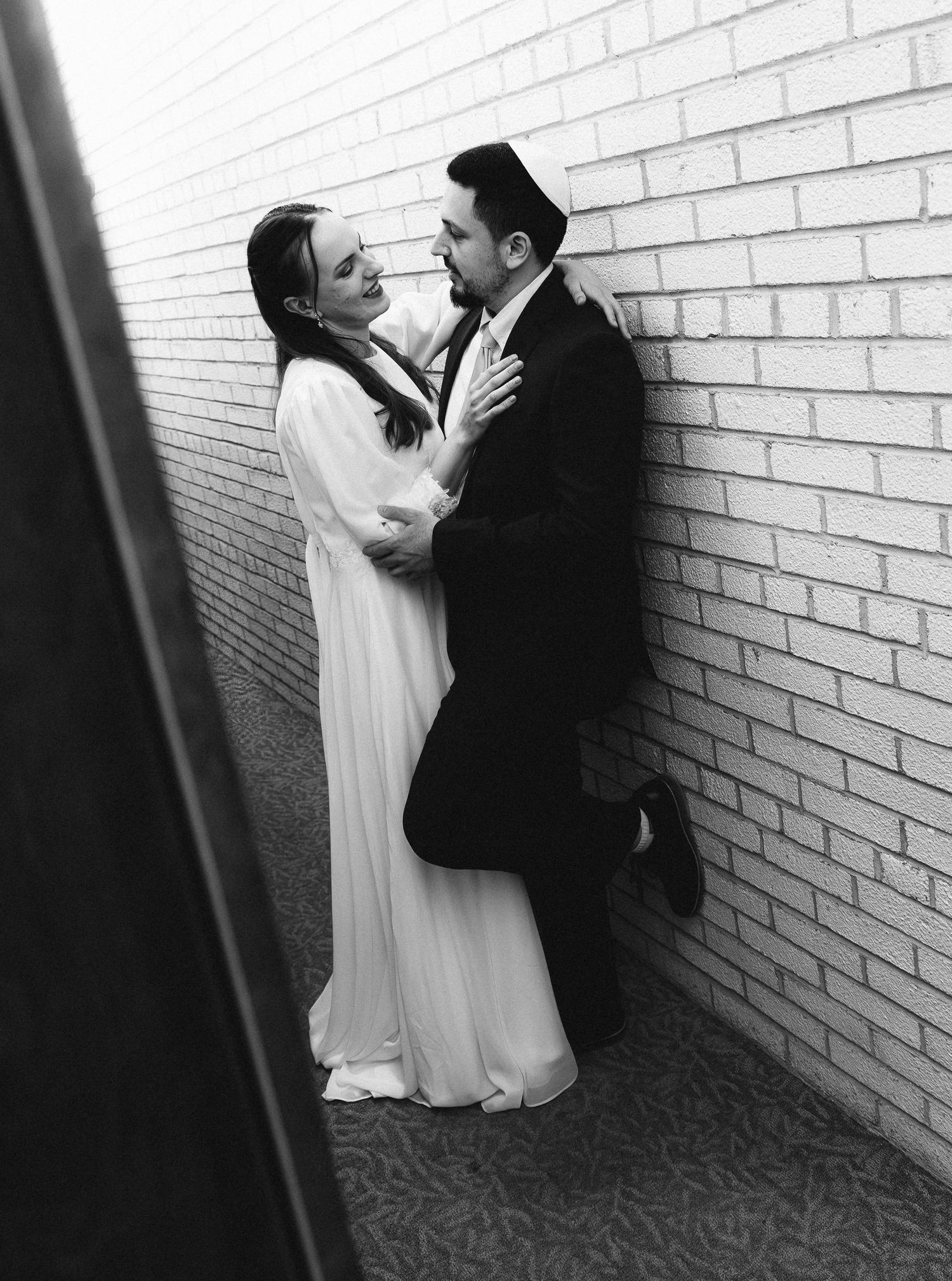Black and white photo of a bride and groom sharing a romantic moment against a brick wall in wedding attire.