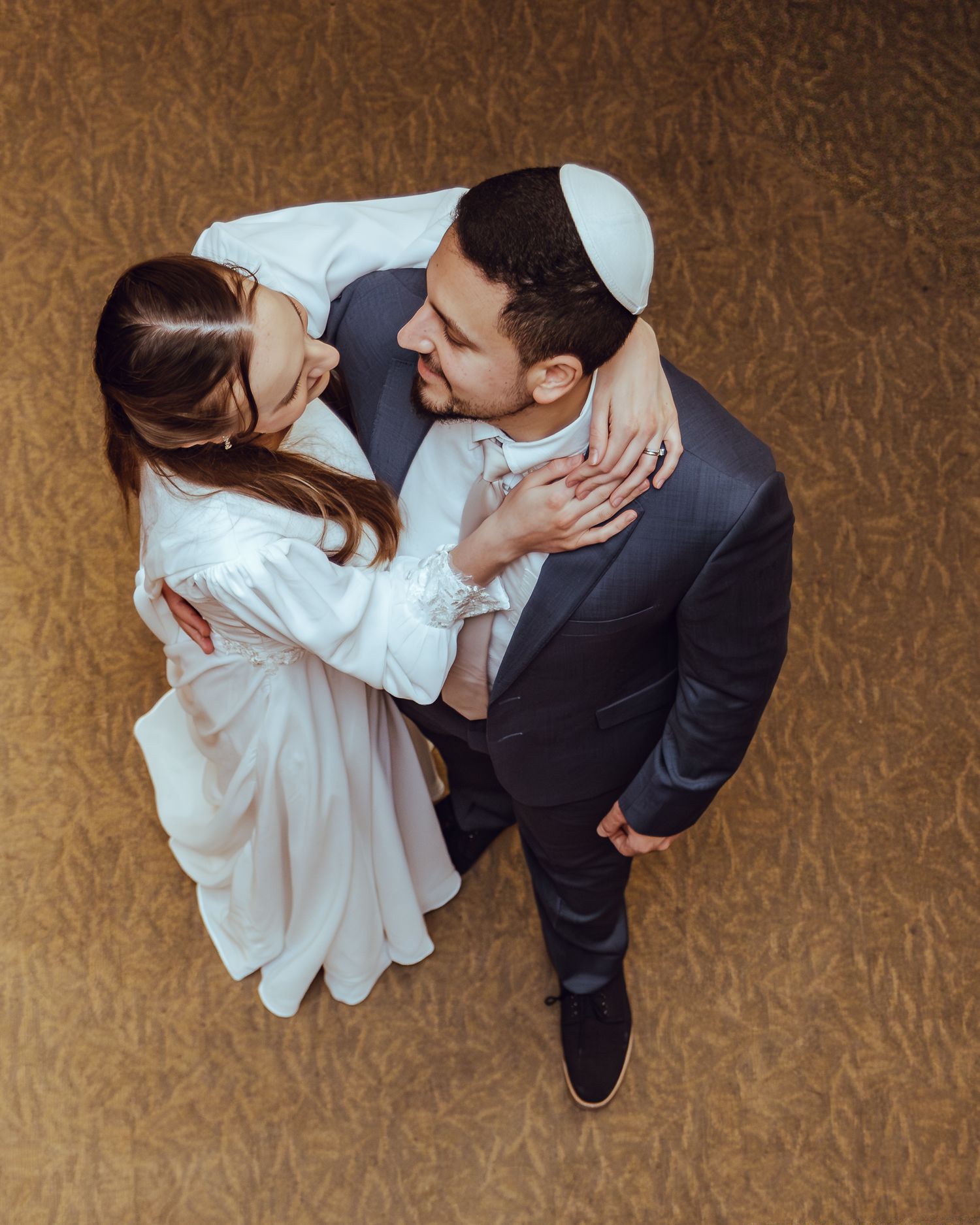 Aerial view of a Jewish wedding couple embracing, groom in suit and kippah, bride in white dress.
