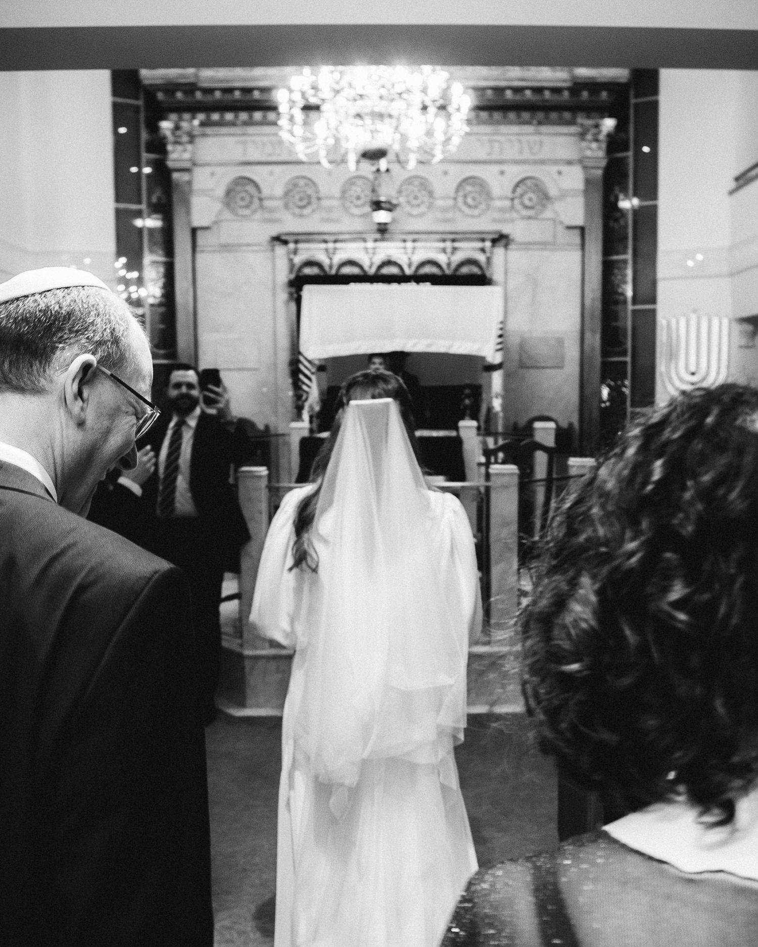 Black and white photo of a bride in veil walking toward the altar in an elegant synagogue during a Jewish wedding ceremony.