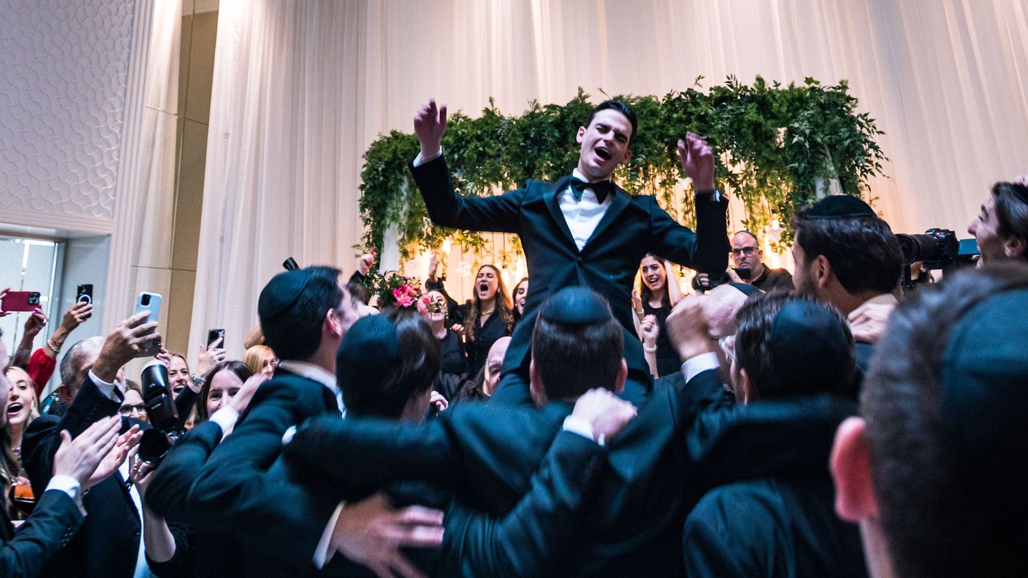 A crowd of wedding guests in formal attire dance and celebrate on a dance floor at an indoor reception.