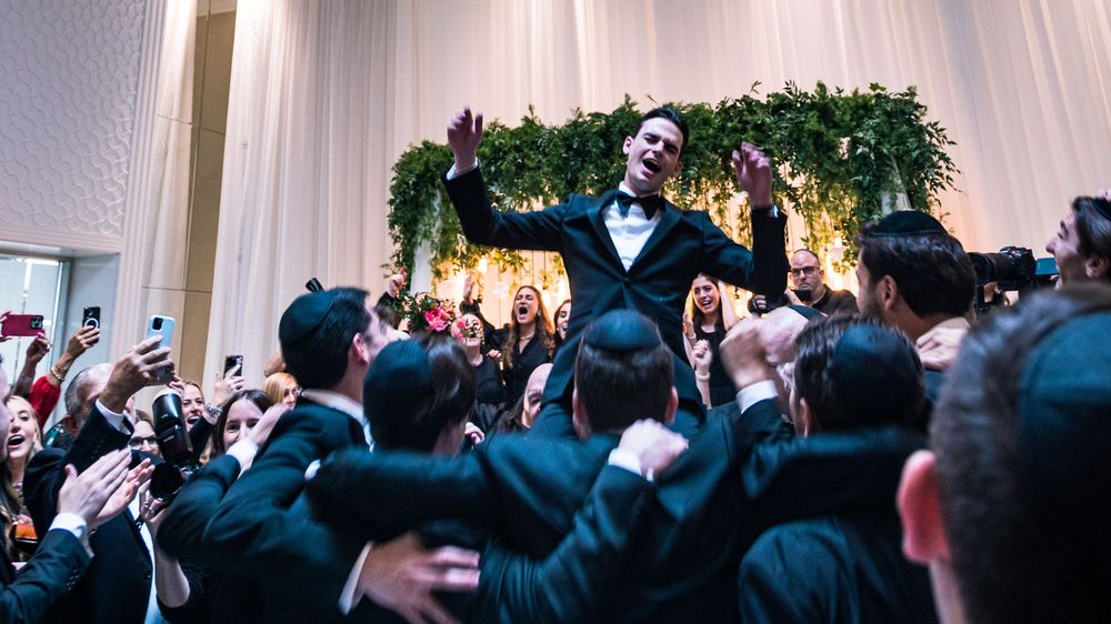 A crowd of wedding guests in formal attire dance and celebrate on a dance floor at an indoor reception.
