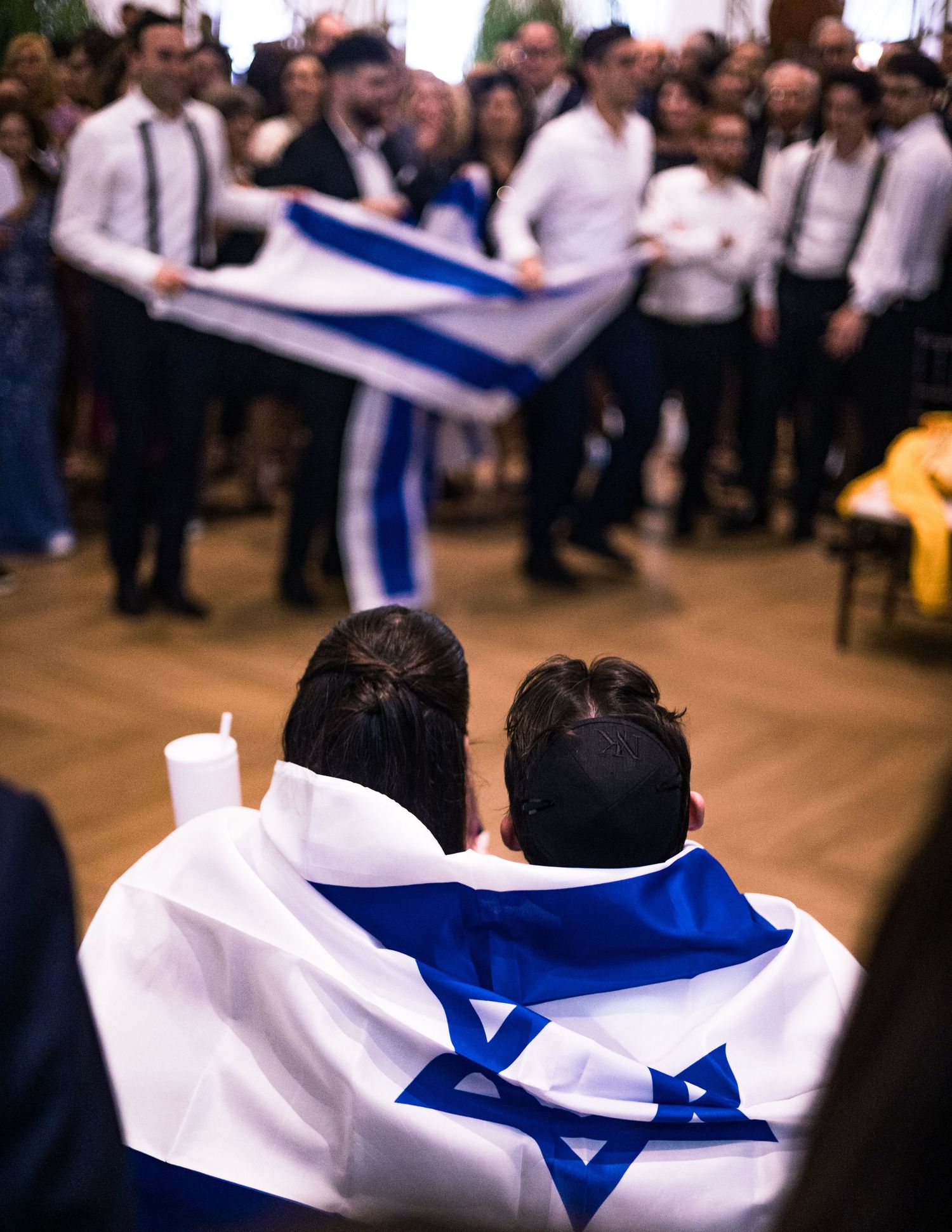 People draped in Israeli flags gather at an indoor celebration event with flags being waved in the background.
