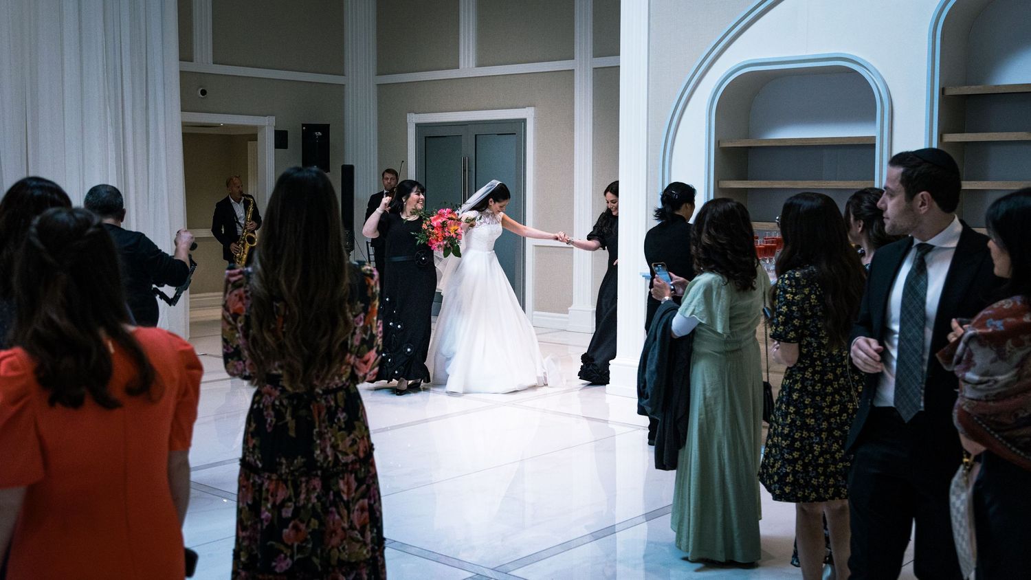 Wedding guests gather around a dance floor as a couple in white and black formal attire dance at an elegant venue.