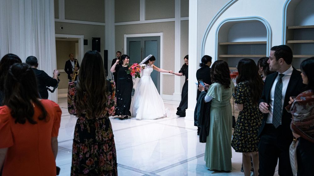 Wedding guests gather around a dance floor as a couple in white and black formal attire dance at an elegant venue.
