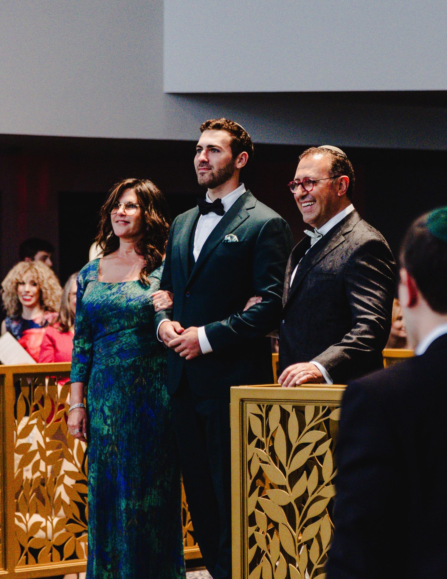 People in formal attire stand together in a line against a dark background with decorative gold panels.