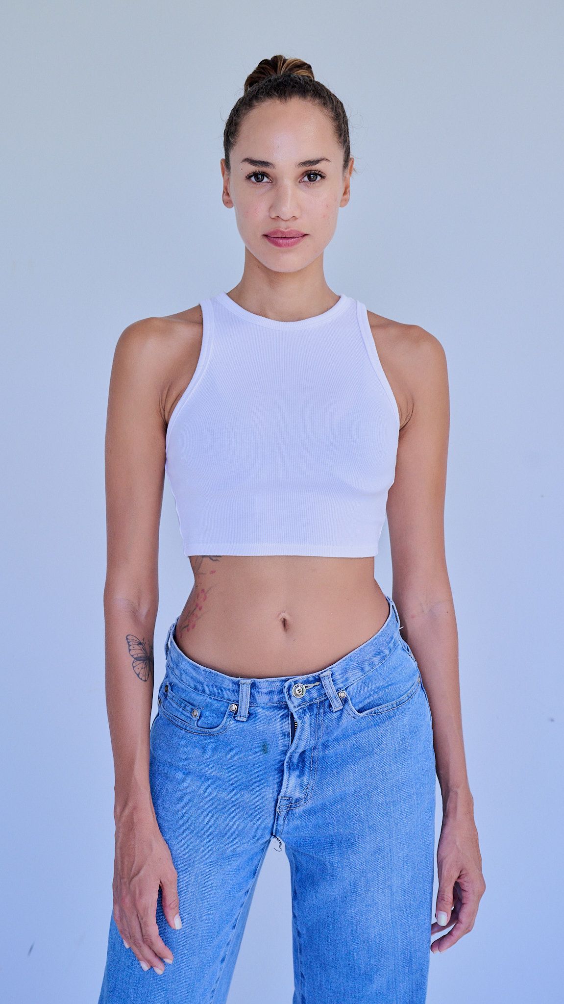 Model wearing a white crop top and light blue high-waisted jeans poses against a light blue background.