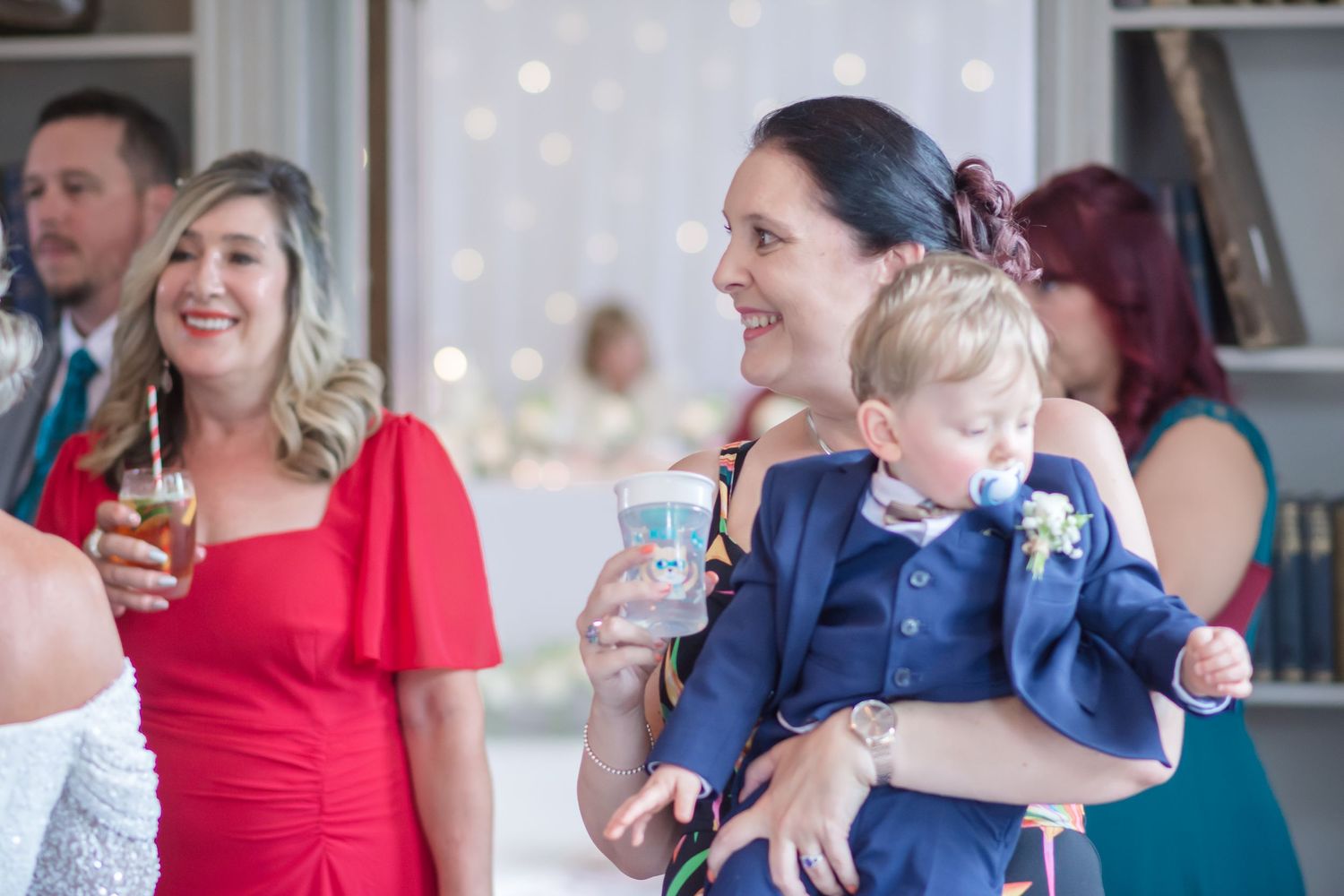 Wedding guests enjoy drinks and laughter at a reception while a young boy in a navy suit rests in arms at DeVere Cranage Estate in Cheshire