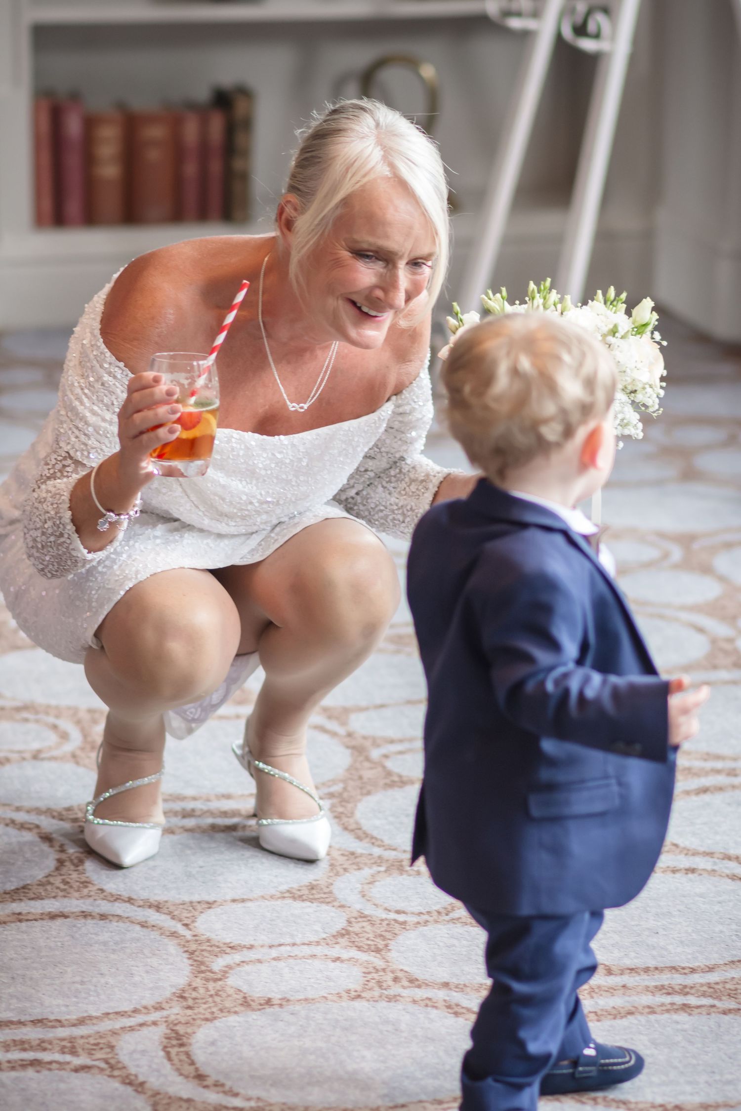 A sweet moment between a woman in a white lace dress and a small child in a navy suit on a patterned carpet.