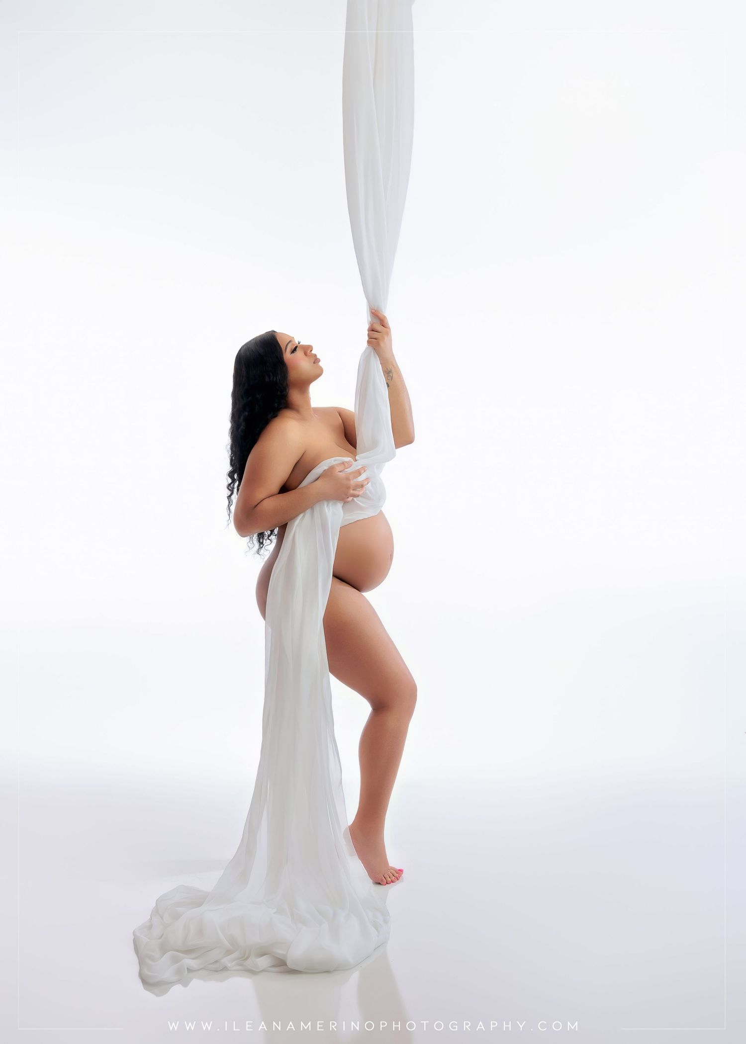 Pregnant woman poses with flowing white fabric draped around her body in bright white studio.