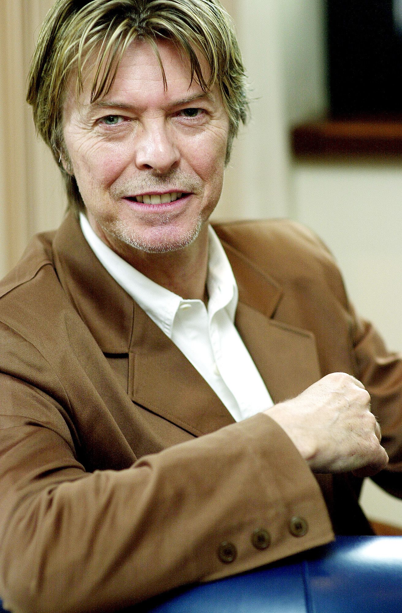 A person wearing a brown blazer and white shirt smiles warmly while sitting in a relaxed pose.