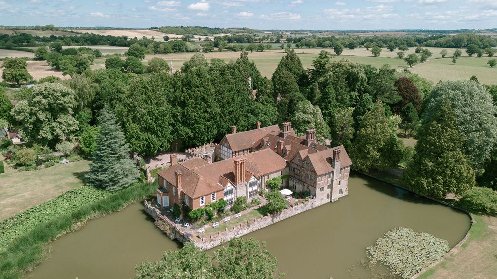 Aerial panoramic view of a Cotswold countryside estate with red brick manor house, gardens and surrounding green fields.