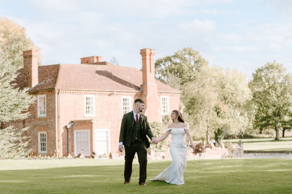 A Worcestershire couple in wedding attire walks on a green lawn in front of Birtsmorton Court on a sunny day.