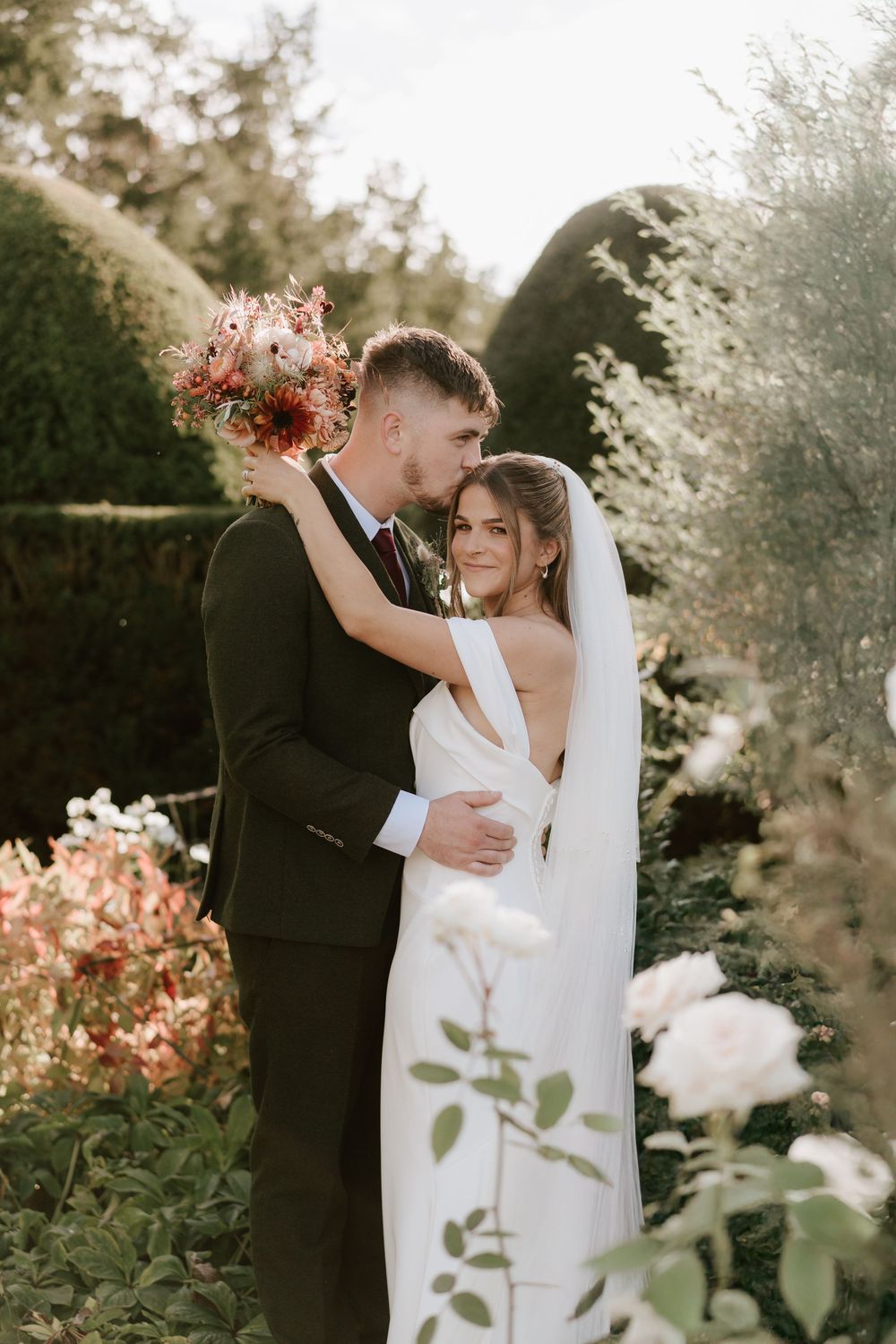 A romantic garden wedding photo with a couple embracing among blooming flowers and greenery at sunset at Birtsmorton Court