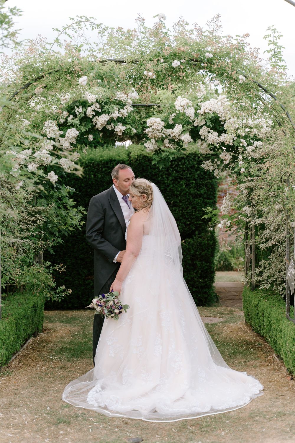 Romantic wedding couple embraces in a lush garden setting with white floral archways and greenery.