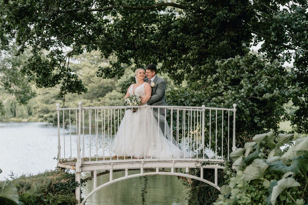 A romantic wedding couple stands on a white arched bridge surrounded by lush greenery in a garden setting.