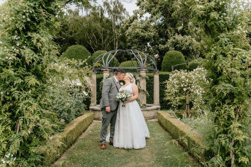 A garden wedding ceremony setup with lush greenery archways and white chairs along a stone pathway.