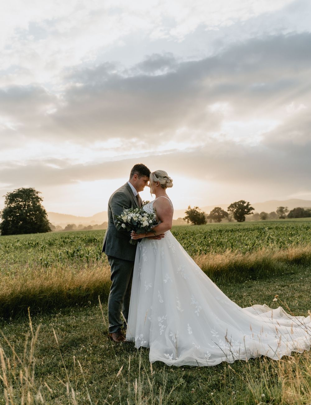 A romantic couple embraces in a golden field at sunset while wearing formal wedding attire. At Birtsmorton Court in Worcestershire.
