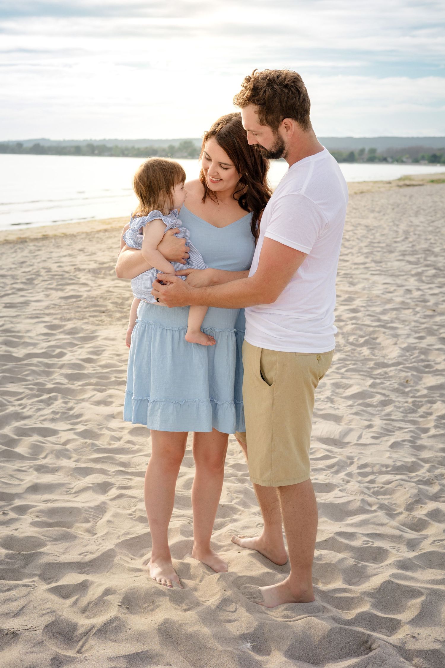 Family wearing coordinated blue and white outfits embracing together on a sandy beach at sunset.