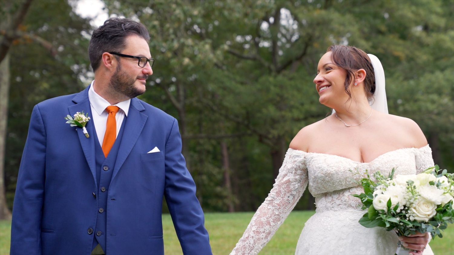 A couple in wedding attire share a tender moment outdoors, with the bride wearing a white lace gown and groom in navy suit.