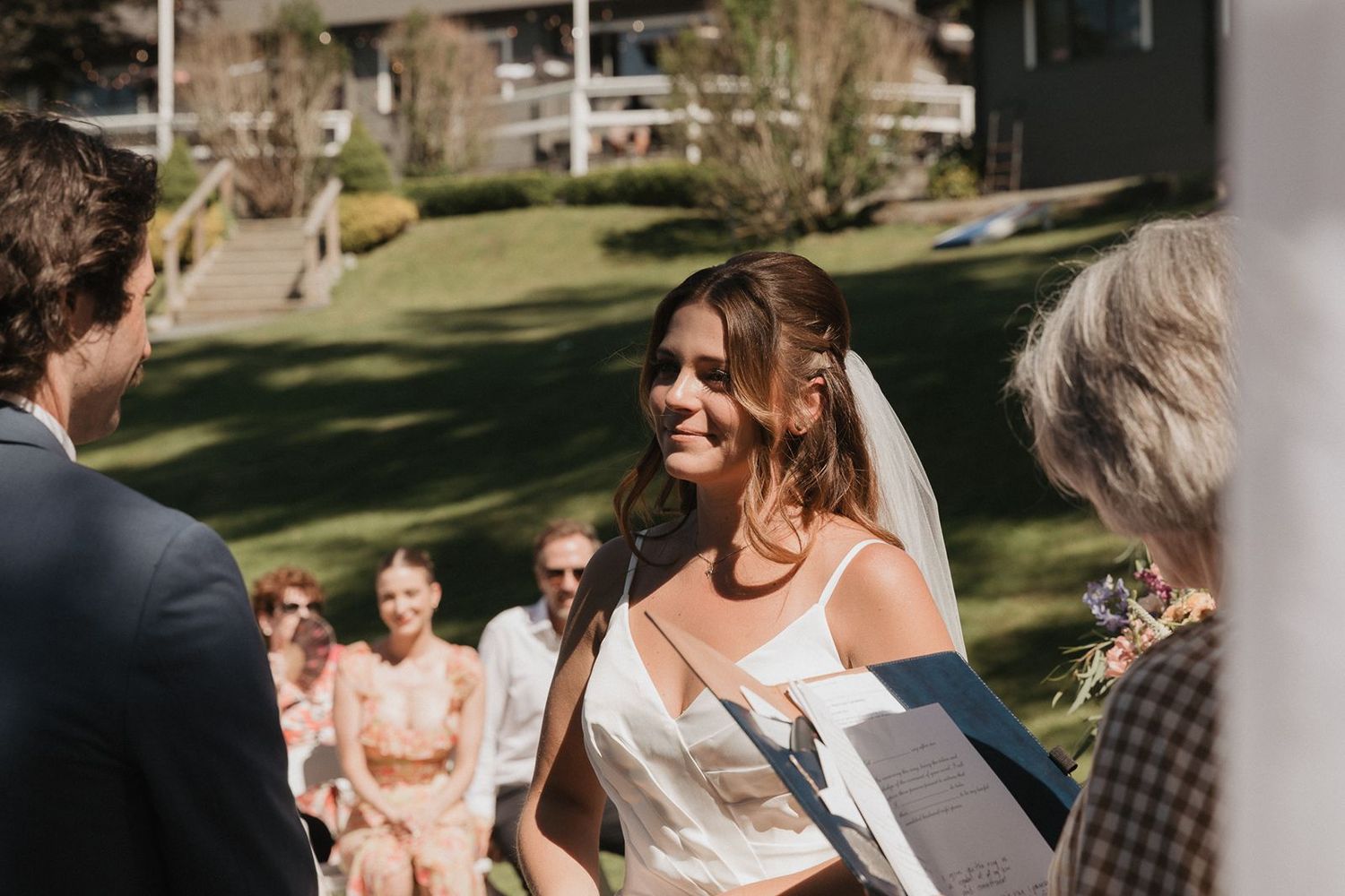 Radiant bride in white veil smiles joyfully at groom during sunny outdoor wedding ceremony.