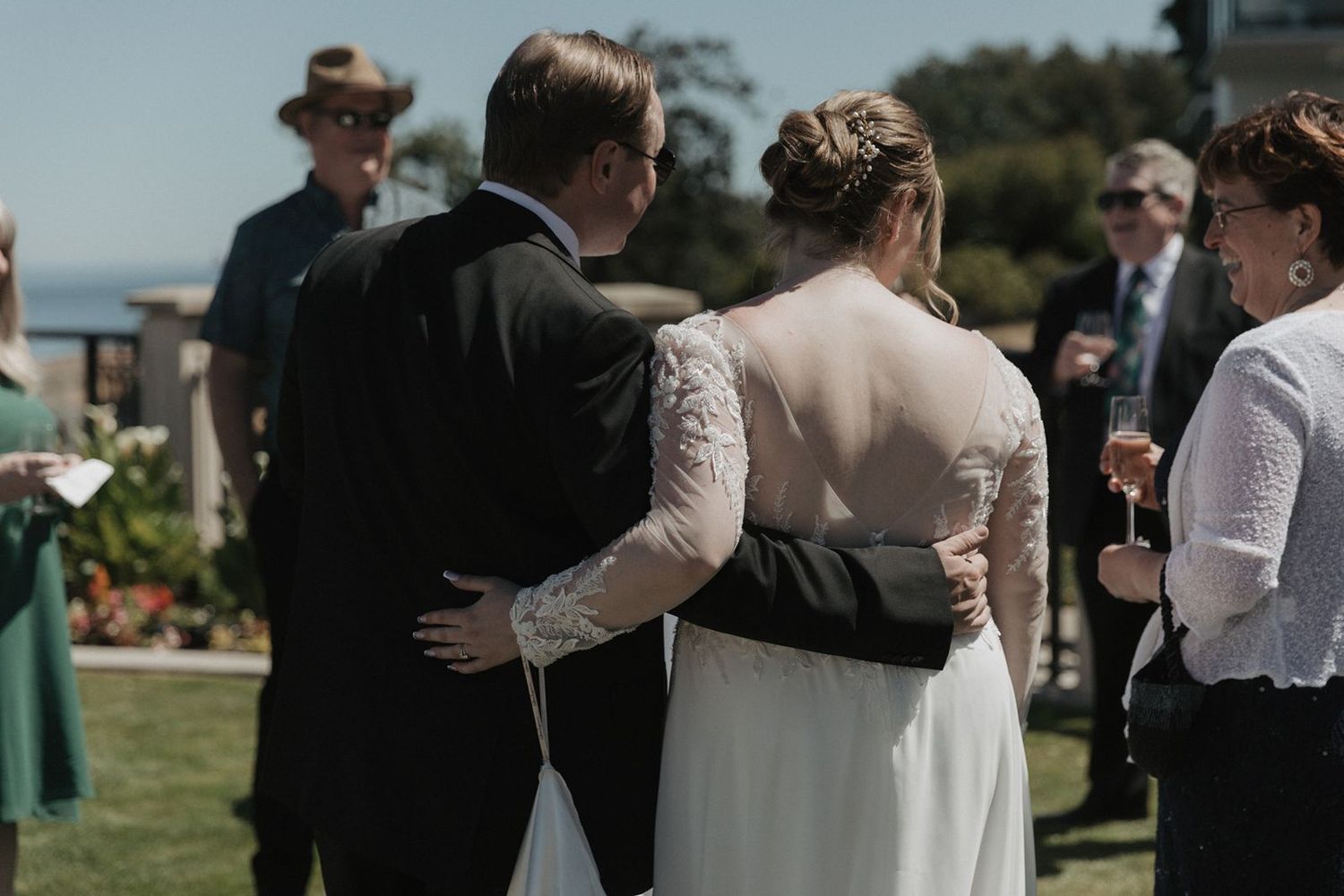 Newlywed couple embraces with arms around each other surrounded by wedding guests on a sunny outdoor lawn.