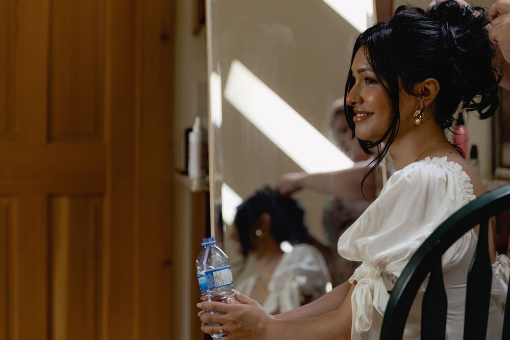 Smiling bride in white dress holding water bottle with dark updo hairstyle and gold earrings.