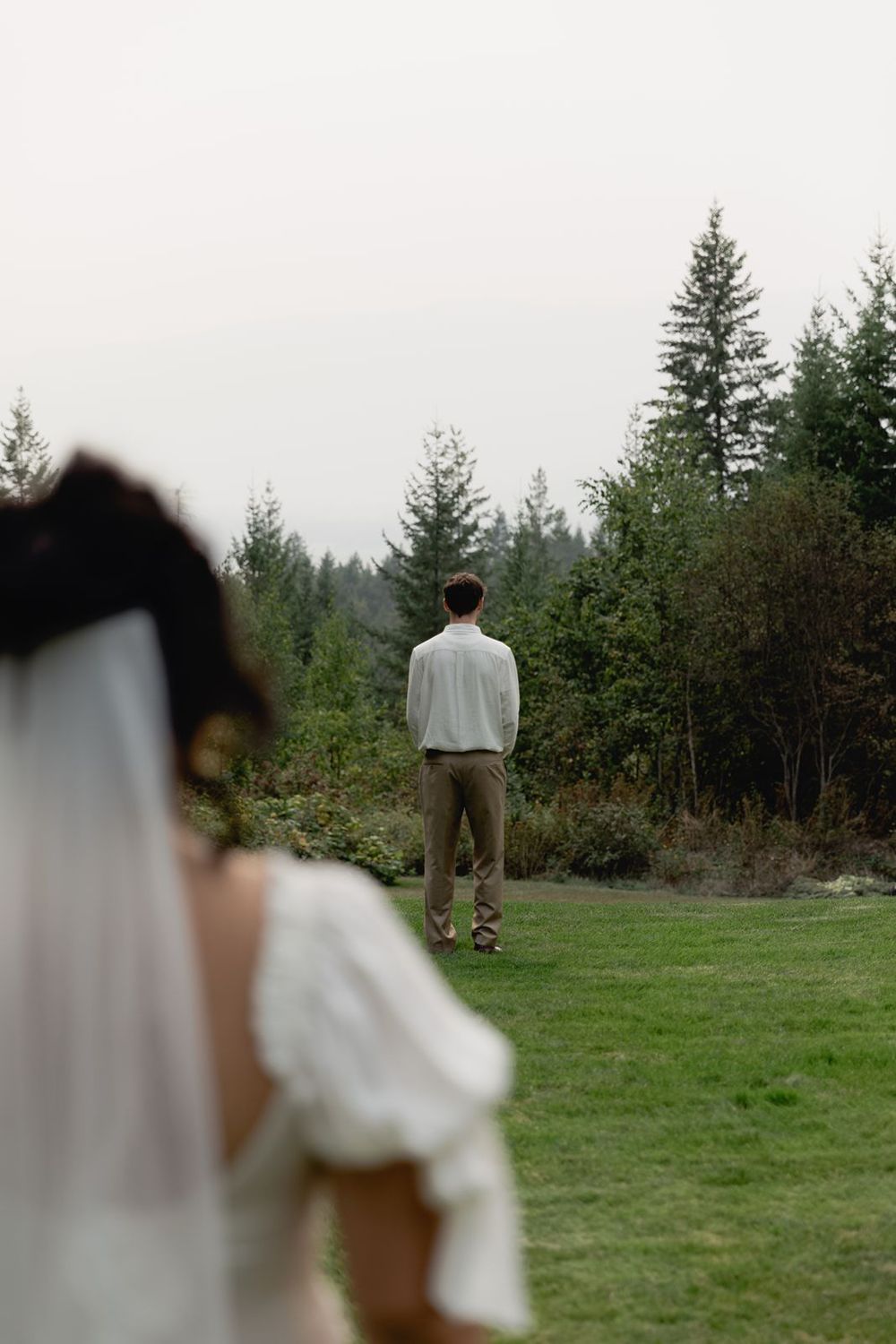 Groom in white shirt and tan pants standing on green lawn facing pine trees as bride approaches.