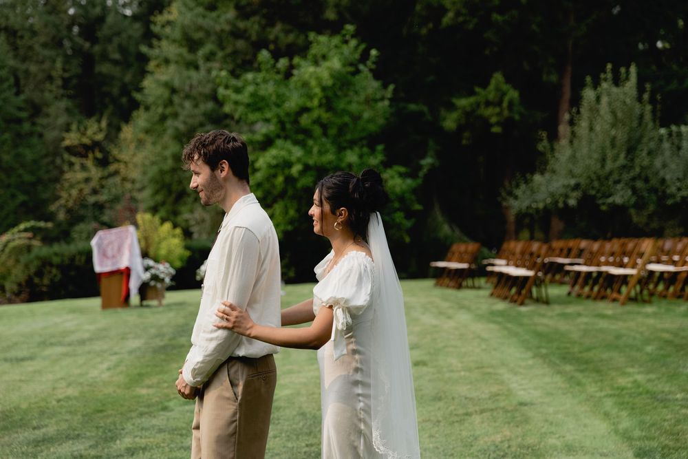 Bride in white gown with veil and groom in white shirt share first look moment on green lawn.